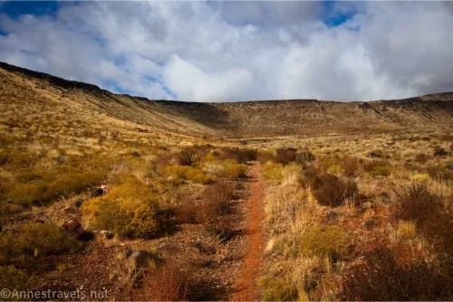 A red trail between desert scrub and clouds in the sky beyond hills