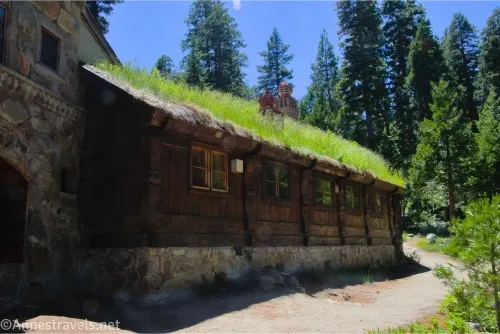A wood and stone building with grass on the roof beside a trail and trees