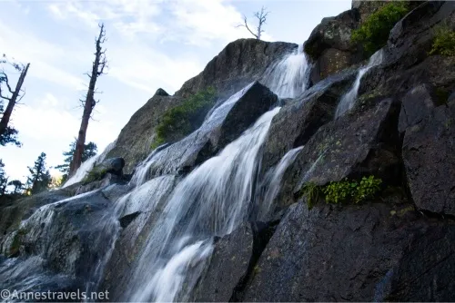 Looking up at a waterfall tumbling over rocks, dead trees, and clouds in the sky