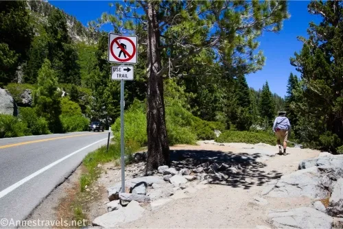 A road, a sign with a pedestrian crossed out, and a hiker on a trail between trees
