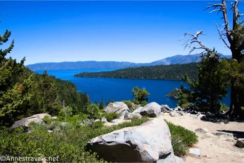 A lake and mountains are visible beyond nearby rocks, bushes, and trees