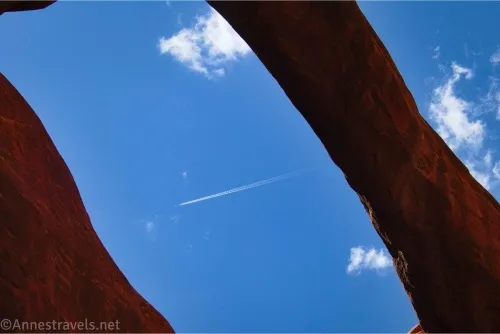 Blue sky, clouds, and an airplane trail beyond an arch