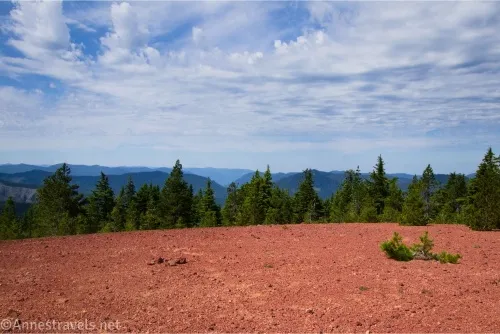 Clouds over green hills, a green pine forest, and red gravel on the ground 