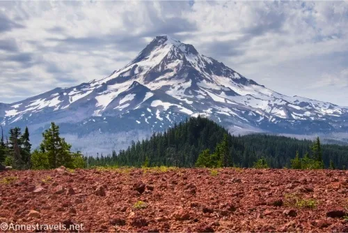 Clouds over a snowy volcano with green trees in the middle ground and red gravel in the foreground 
