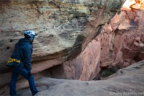 A man with a yellow waist pack and a bicycle helmet stands beside a huge cliff wall and looks down into a deep canyon 