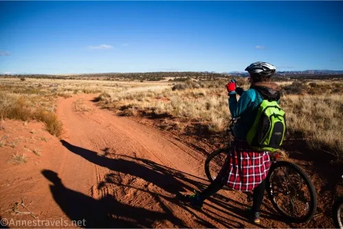A woman in a plaid skirt, a green backpack, and a bicycle helmet on a dirt road in a desert meadow
