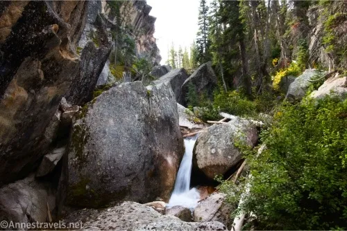 A narrow waterfall tumbles between large rocks bordered by trees and bushes