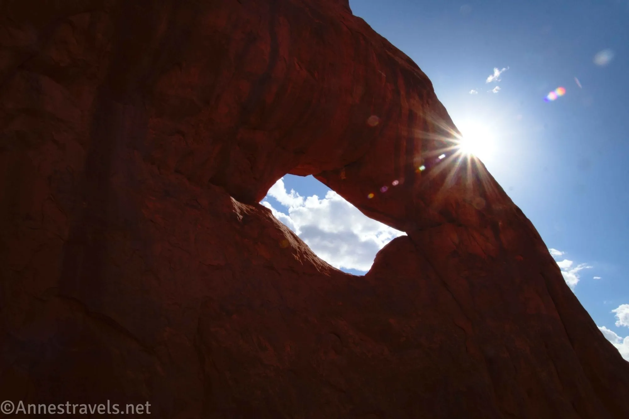A sunstar and an arch against a blue sky with a few puffy white clouds 