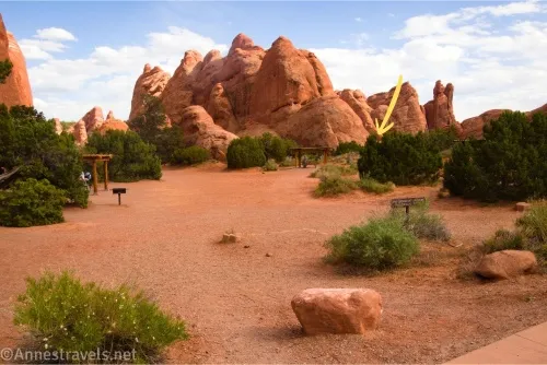 Red rock formations against a blue sky with clouds across an open space surrounded by green bushes 
