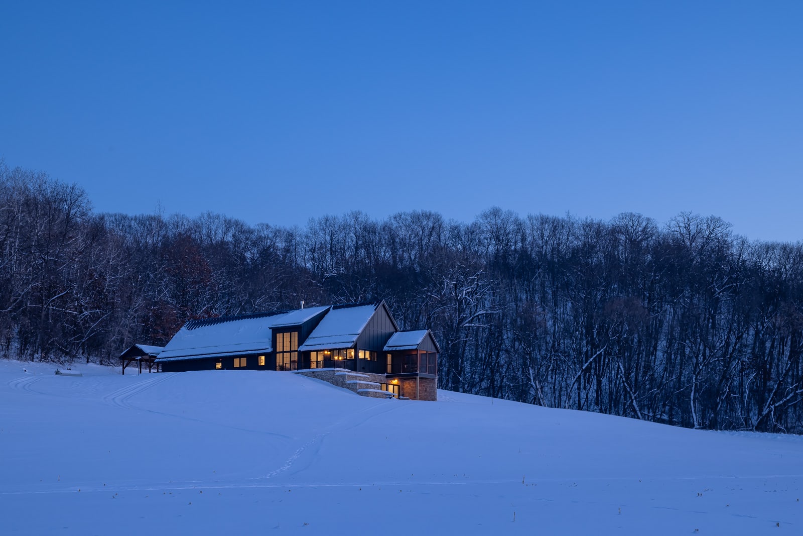 A wide angle, exterior view of the back of the entire Flower Valley Homestead residence just after sun down in the winter. The interior windows are lit up. Black siding covers the majority of the house in view, but the lower level walkout is covered in limestone. The roof is covered in snow, and so is the surrounding landscape.