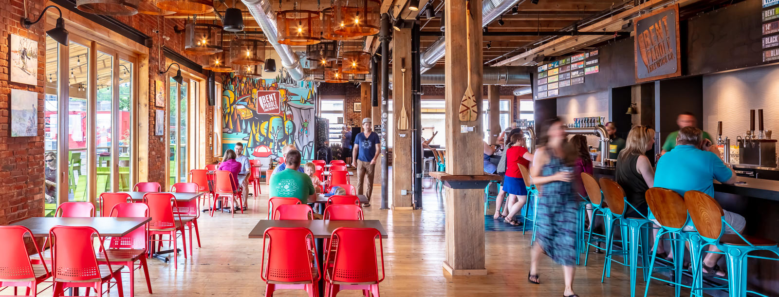 View of the Bent Paddle Brewing taproom, a renovated industrial warehouse. There is table seating with red chairs and a bar with blue barstools. People are sitting and drinking.