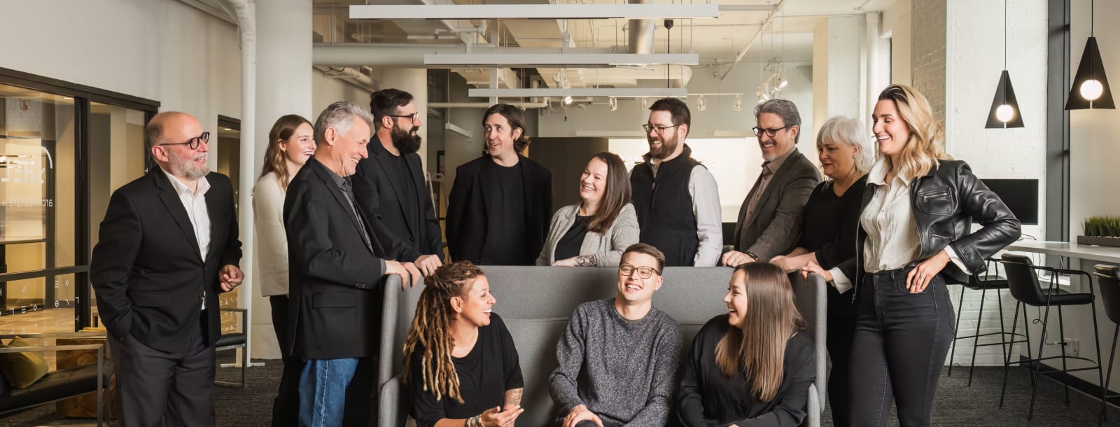 Members of the Shelter Architecture and Interior Design team in their Minneapolis, MN studio. Three people are sitting on a grey couch with more people behind them surrounding it.