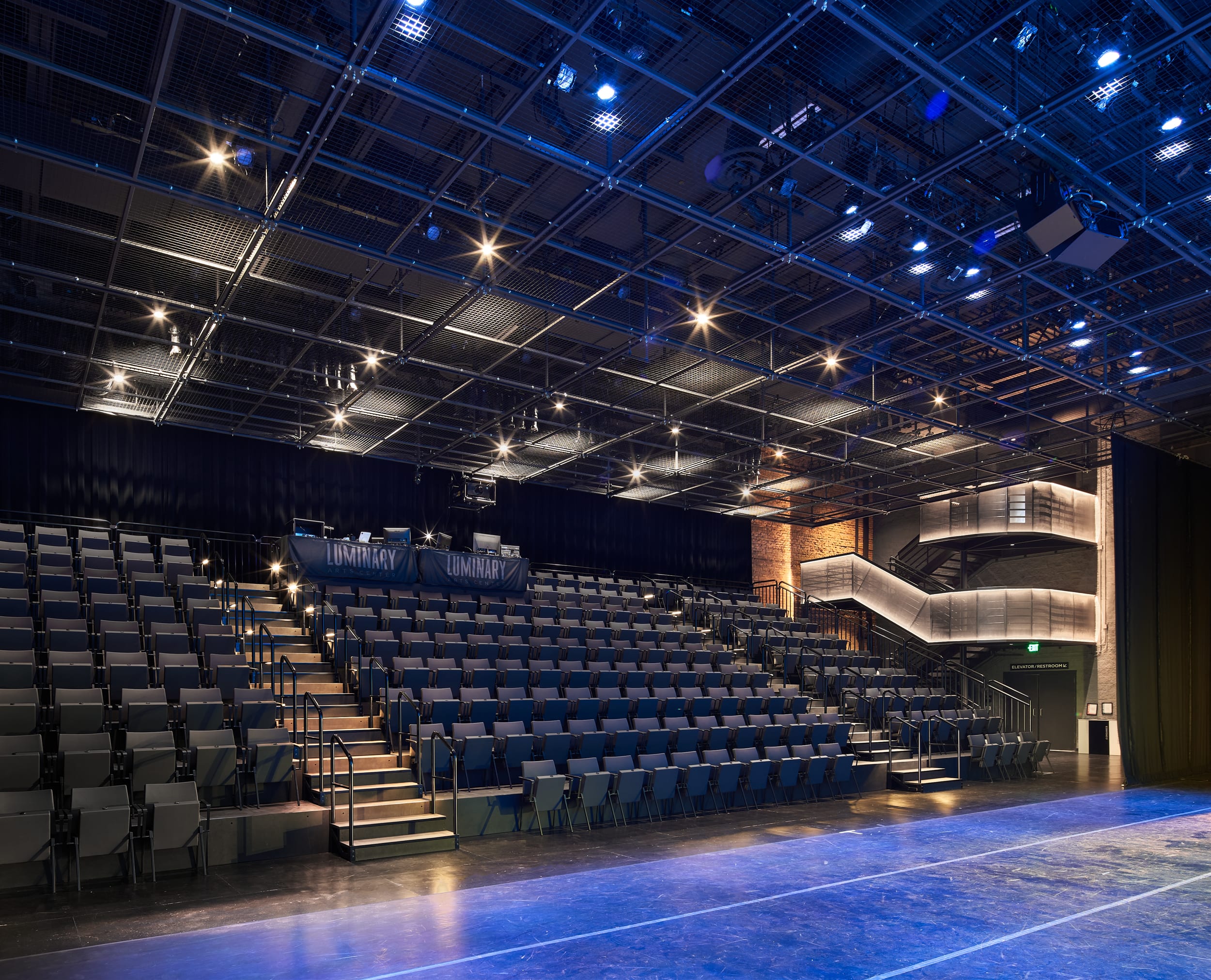 The theater seating, illuminated main staircase, and tension wire lighting grid suspended from the ceiling in the Luminary Arts Center performance space in Minneapolis, MN.