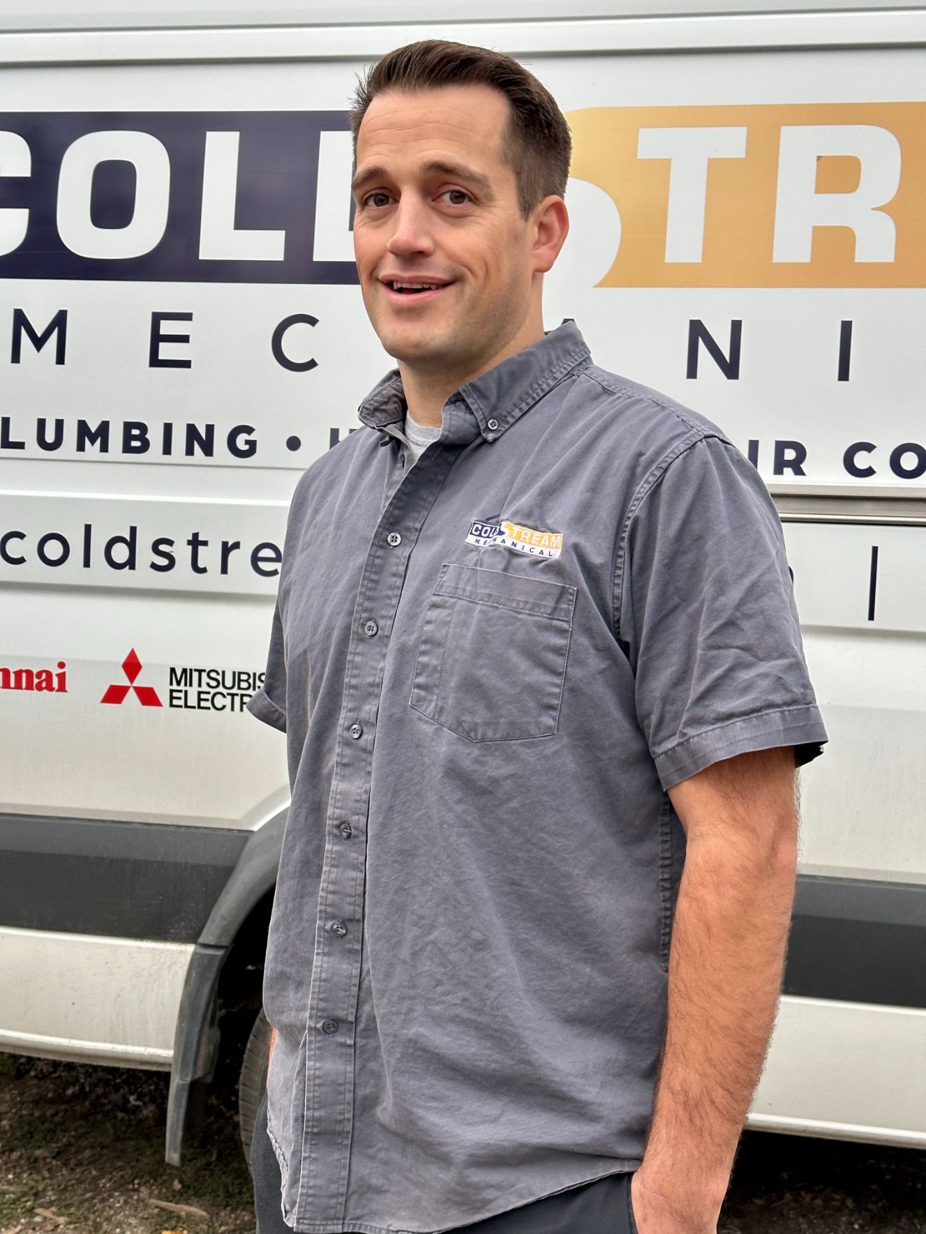 david hamm, owner of coldstream mechanical, stands smiling in front of a company van wearing a grey company shirt with his hands in his pockets