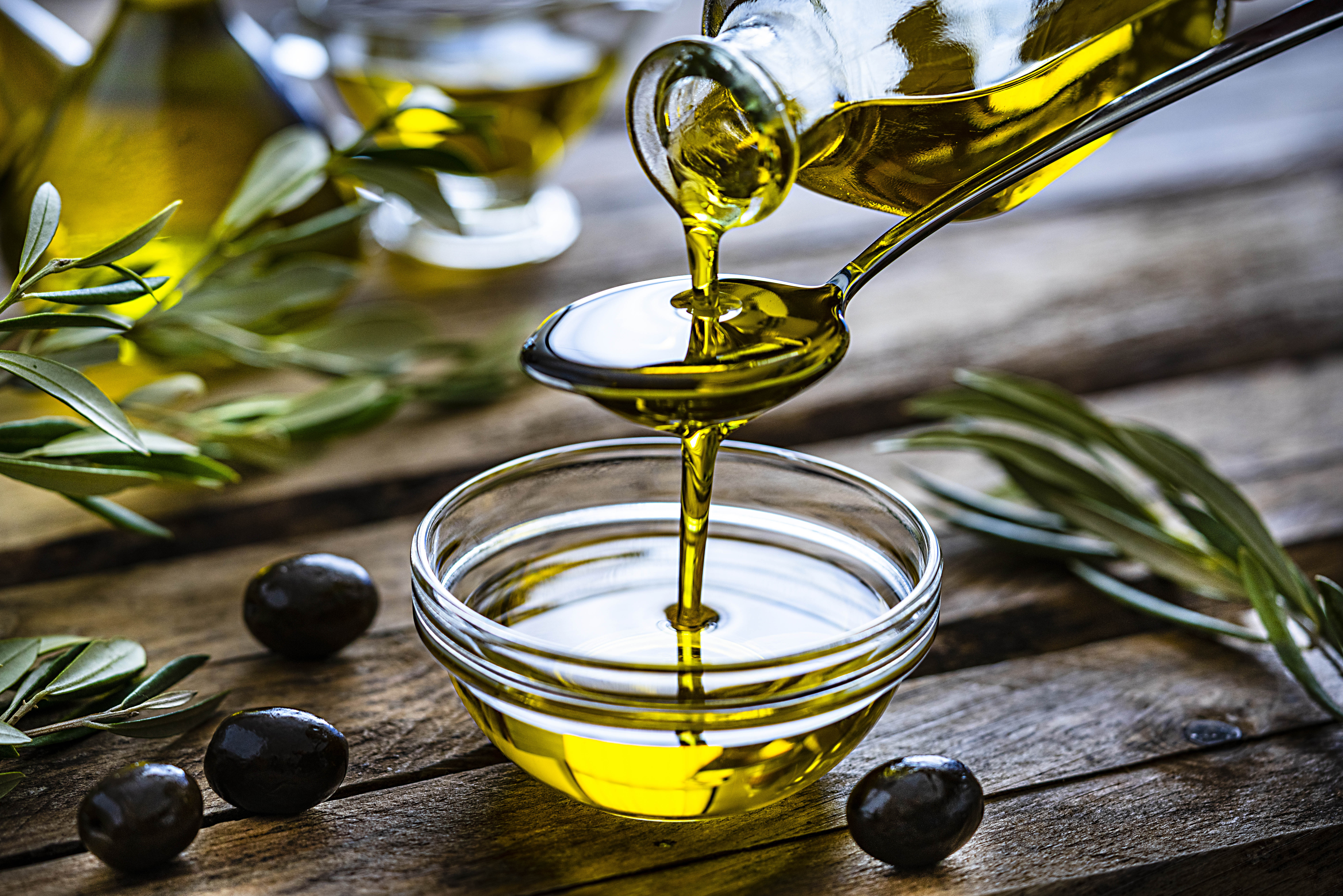 Pouring extra virgin olive oil from a spoon to a glass container. Olive branches comes from the left and right. Some olive oil bottles are out of focus at background. Black olives are on the table and complete the composition. The composition is on a rustic wooden kitchen table. Predominant colors are gold, green and brown.