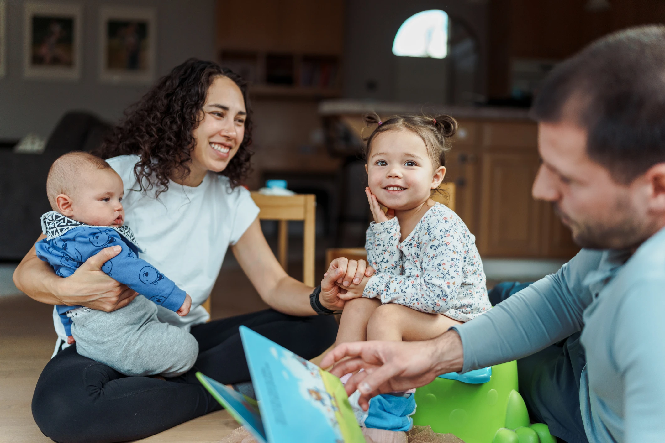 A multiracial woman sits on the floor while holding her baby son and congratulates her toddler daughter for using the potty. The toddler's dad is holding a storybook and providing support and encouragement.