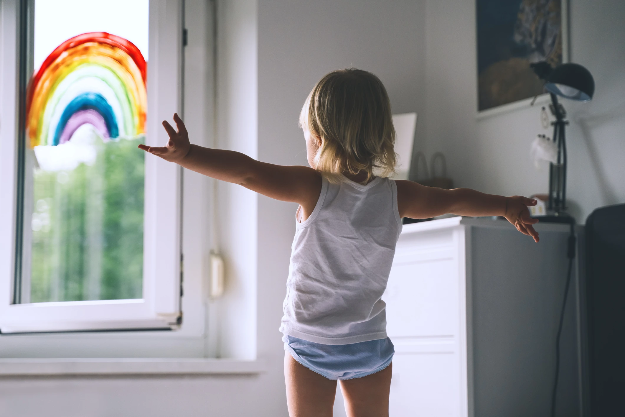 Cute little girl having fun time jumping on bed on background of window with a painted rainbow. Kids leisure and play at home. Image of healthy and happy childhood, creativity, family, joy and fun.