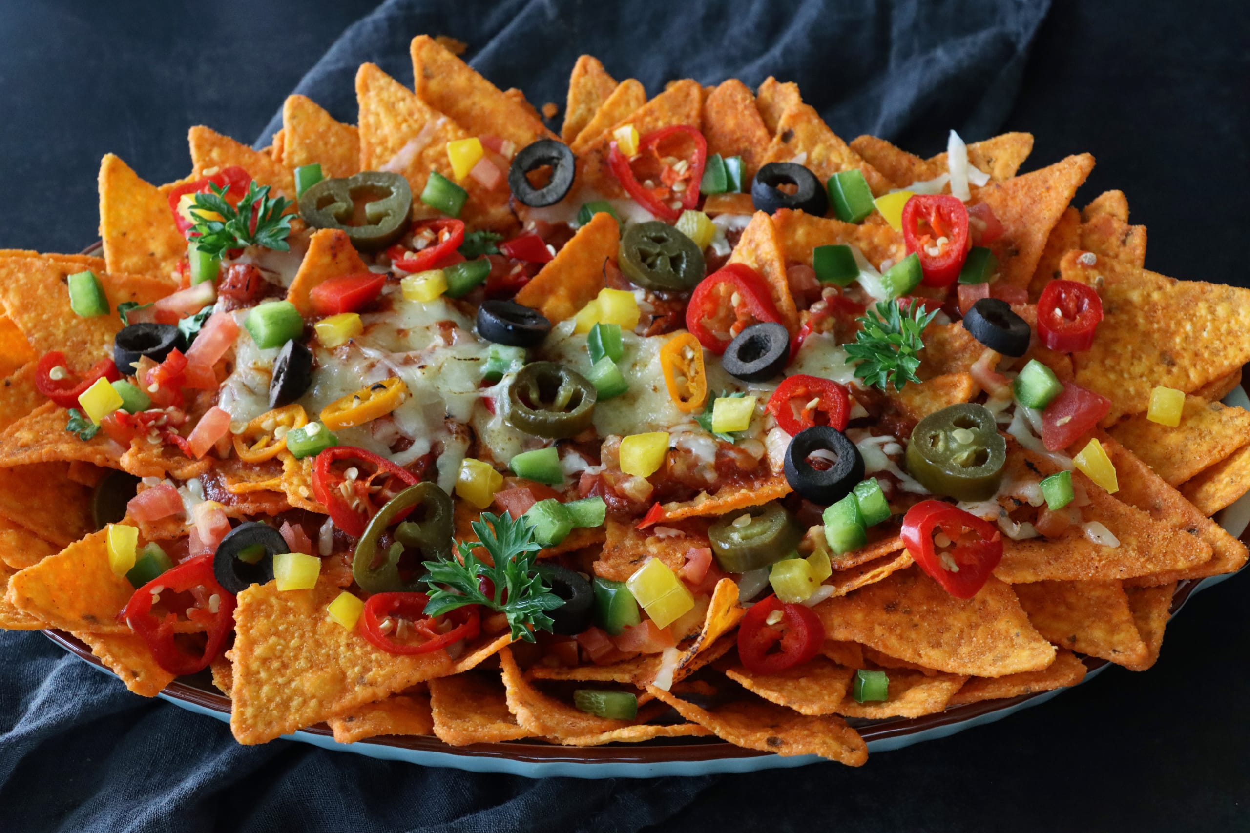 homemade loaded nachos recipe. This snack contains fresh, crunchy tortilla chips covered in melted cheese with cherry tomatoes, black olives and Jalapeno peppers on an oval plate, served with lime wedge, salsa, sauces and dips.