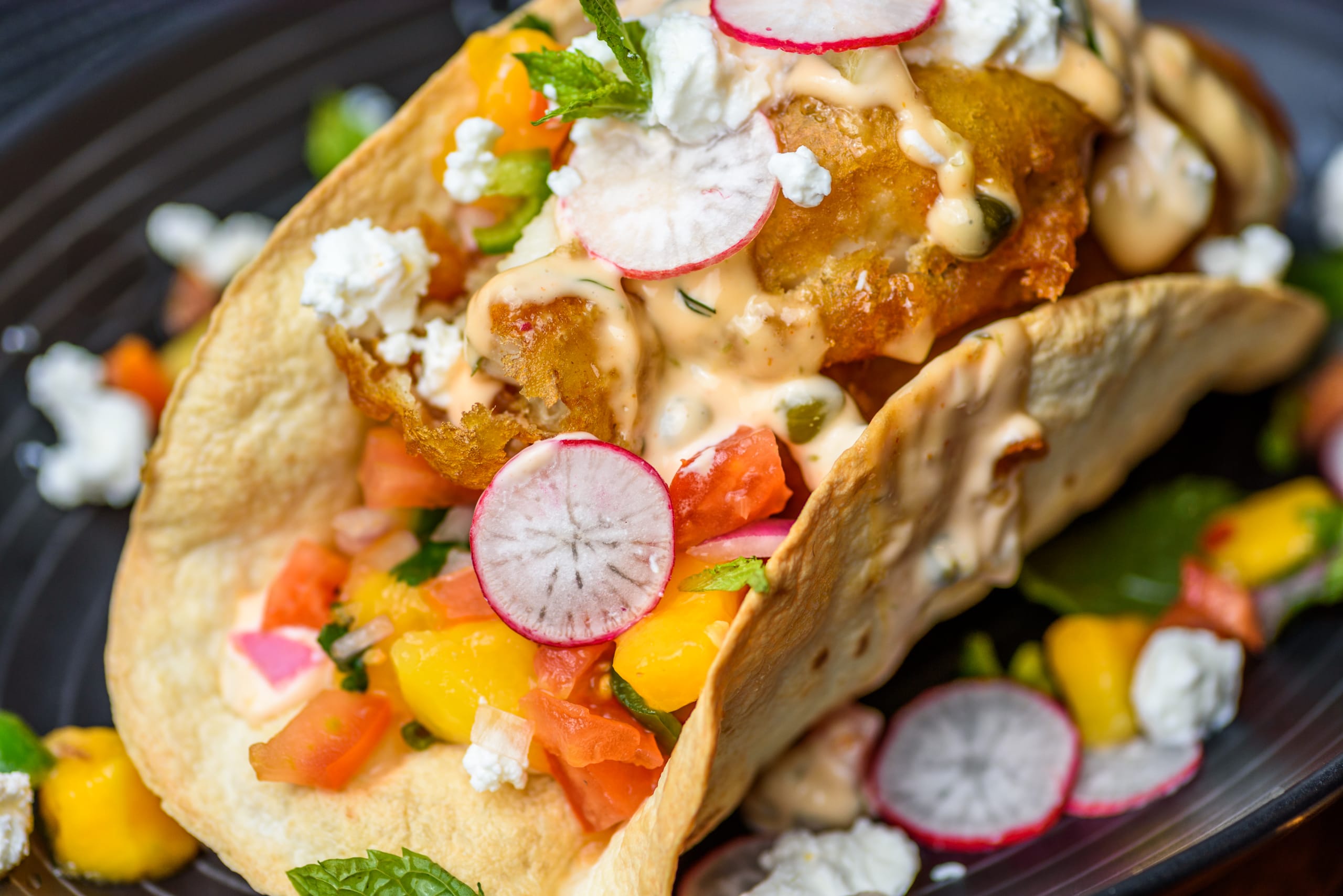 A serving of fish taco with salsa salad, feta cheese, sriracha sauce in a crispy taco shell