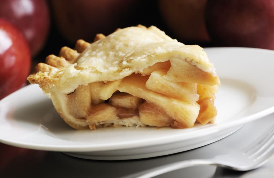 Homemade and thick slice of apple pie in a white plate with apples in background.