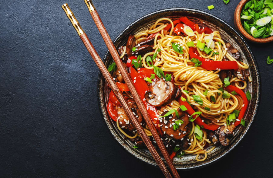 Vegan stir fry egg noodles with vegetables, paprika, mushrooms, chives and sesame seeds in bowl. Asian cuisine dish. Black table background, top view