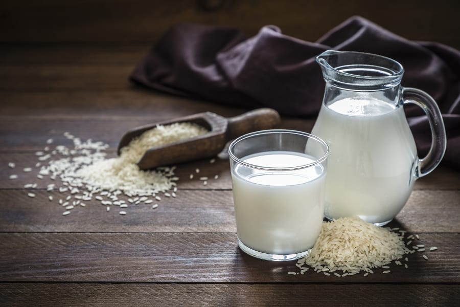 Front view of a jug and a drinking glass full of rice milk. The glass and the jug are at the right side of the image on a rustic wooden table, next to the glass its a heap of rice grains.