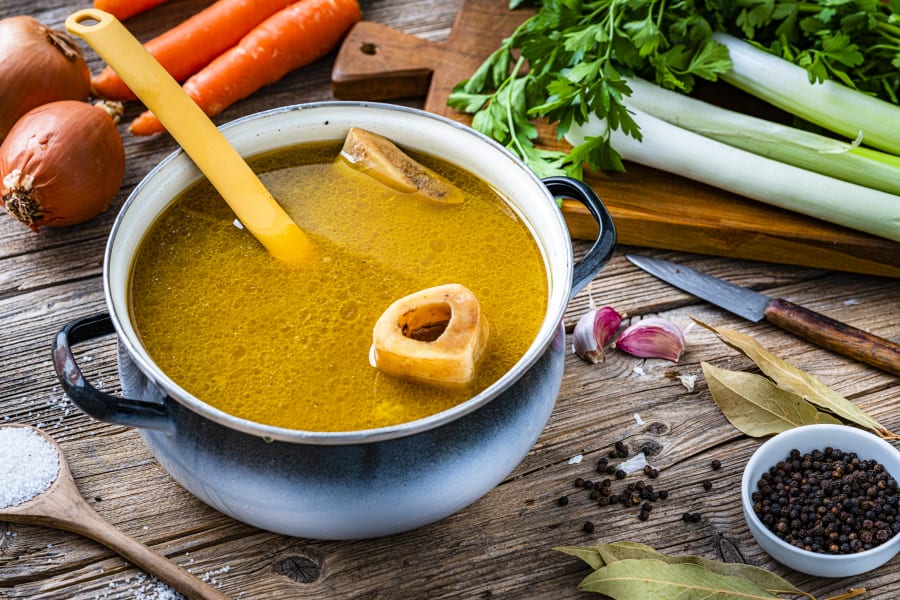 High angle view of a cooking pan filled with homemade bone broth shot on rustic wooden table. Ingredients for cooking bone broth are all around the pan.