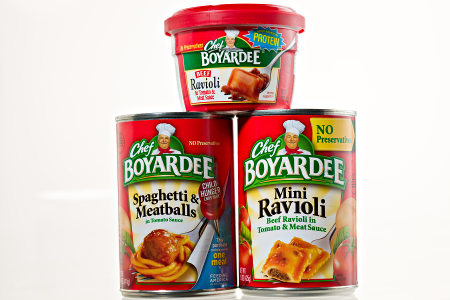 A 7.5 OZ individual plastic container of Chef Boyardee Beef Ravioli sitting on top of two 15 OZ metal cans of Chef Boyardee Mini Ravioli and Spaghetti And Meatballs.