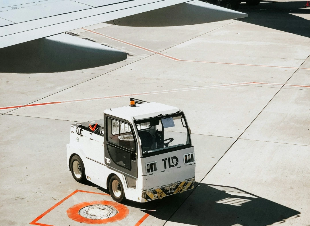 Vehículo tractor TLD en plataforma aeroportuaria bajo el ala de un avión comercial, listo para operaciones de remolque y asistencia en tierra.