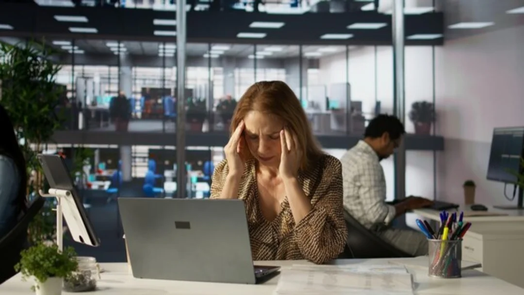 Trabajadora con expresión de estrés frente a un ordenador portátil en una oficina, con las manos en las sienes, ilustrando los riesgos psicosociales y de salud mental en el entorno laboral