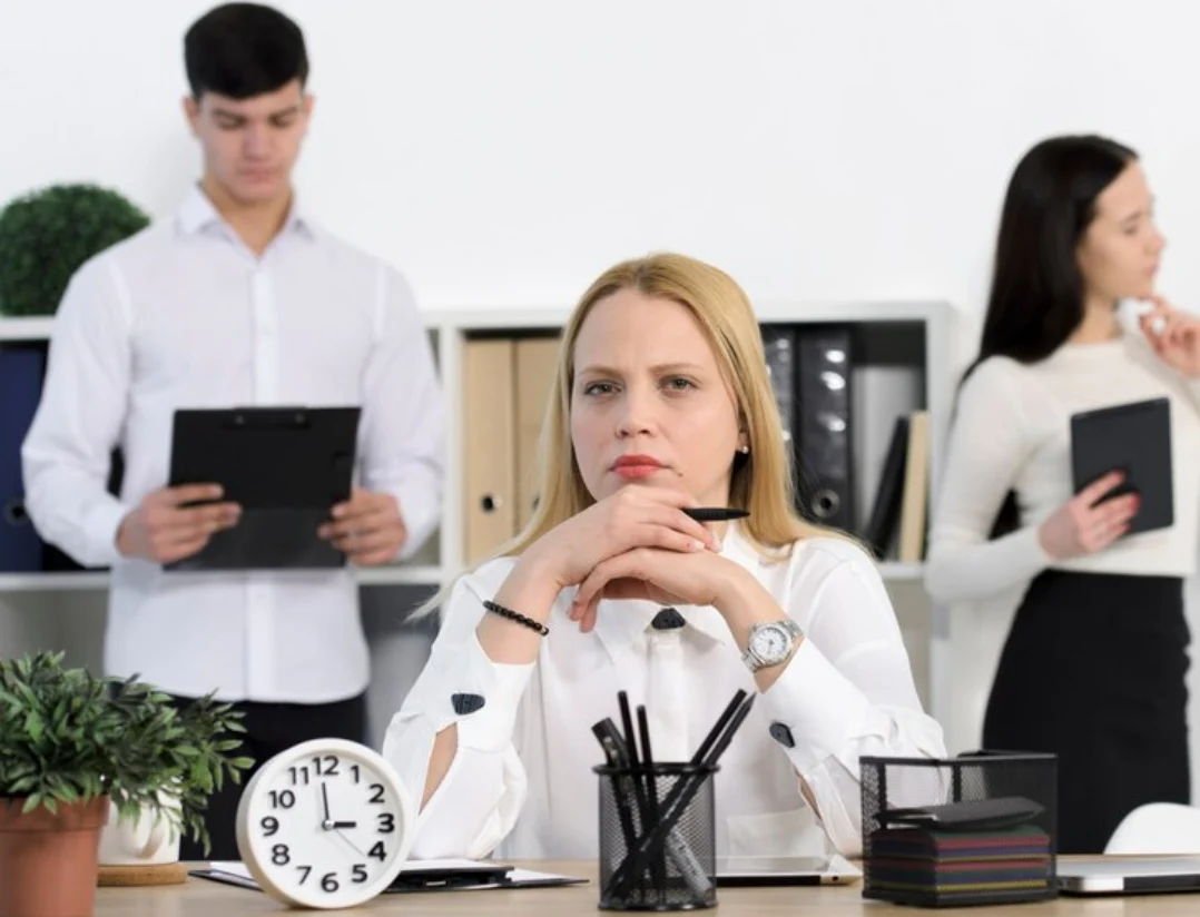 Trabajadora en oficina con expresión reflexiva ante un cambio de funciones, con reloj y documentos sobre la mesa y compañeros al fondo