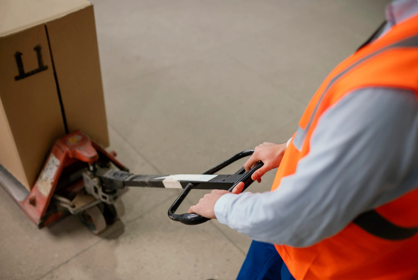 Trabajador con chaleco de seguridad naranja manejando un carro manual de transporte de mercancías en un almacén logístico, escenario habitual de accidentes laborales en empresas subcontratadas