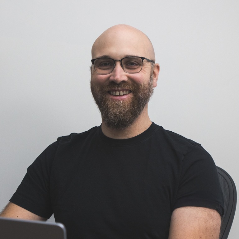 A photo of Adam Wills at his desk, smiling with a laptop