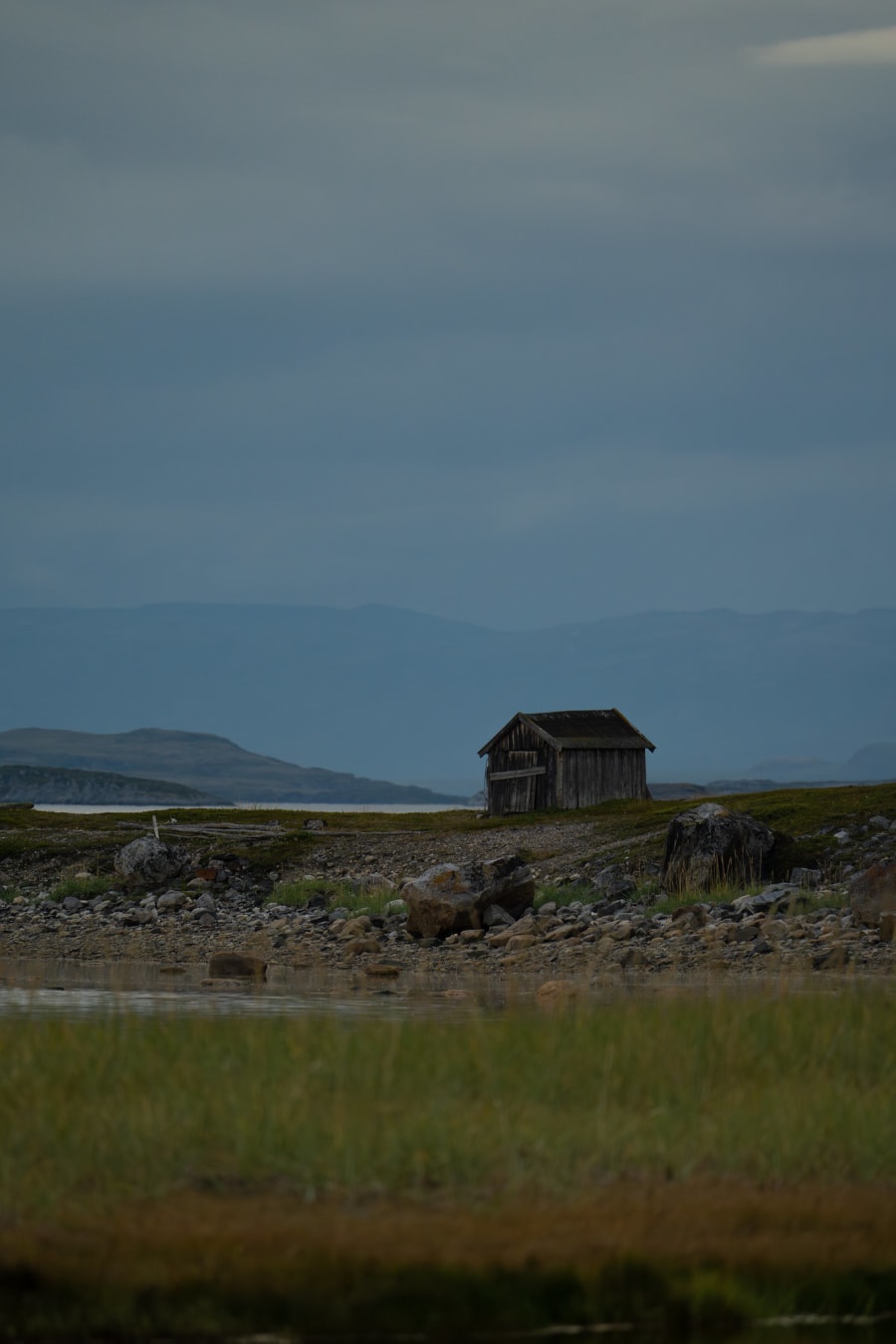 Wooden shed that's falling apart, by a lake surrounded by green grass, and the fjord in the background. The mountains and sky in the background are different shades of blue. The foreground consists of different greens and oranges in the grass.