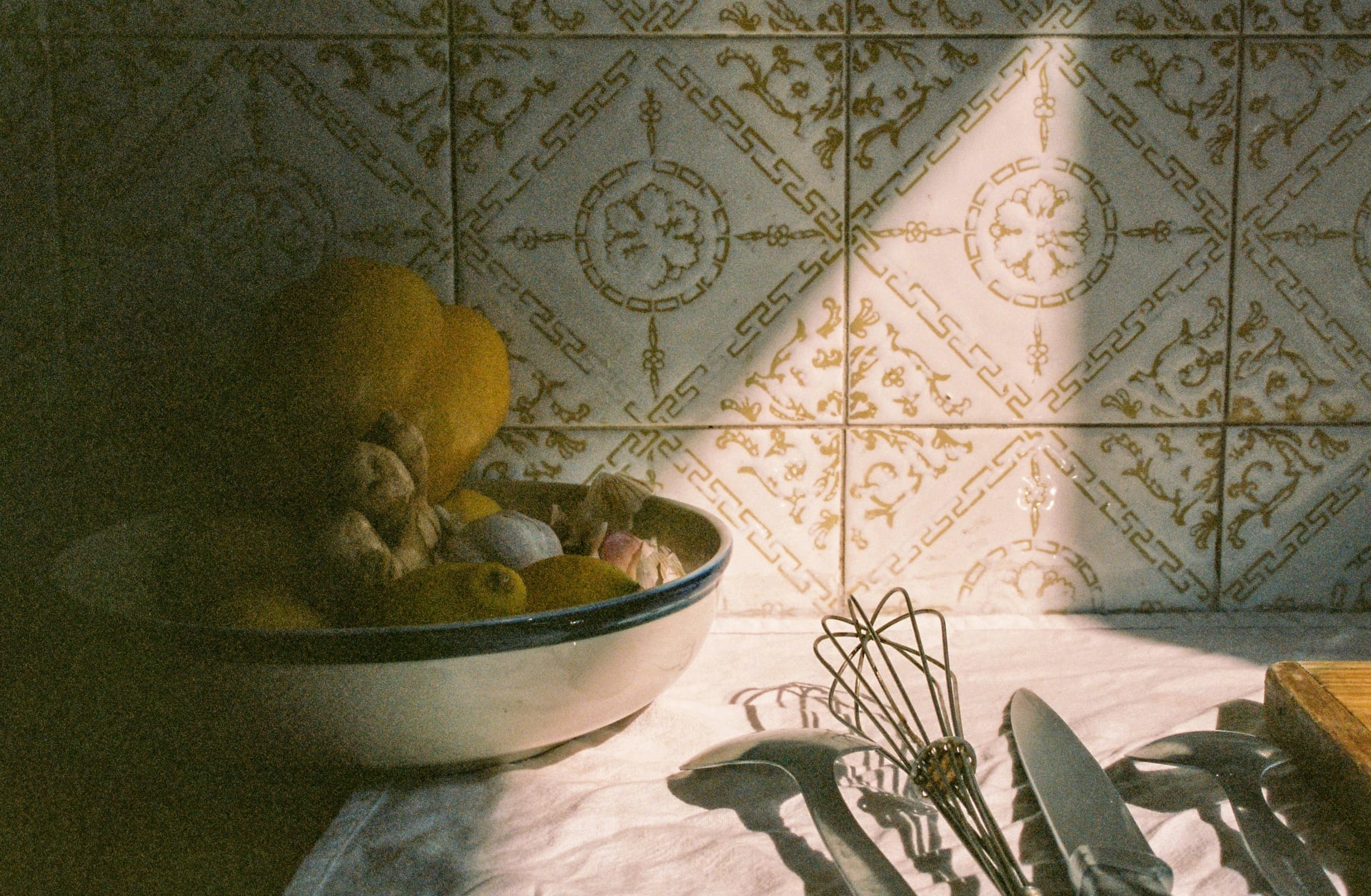 A bowl of fruit, a whisk and cutlery on a kitchen table in late sun against a patterned tiled wall.