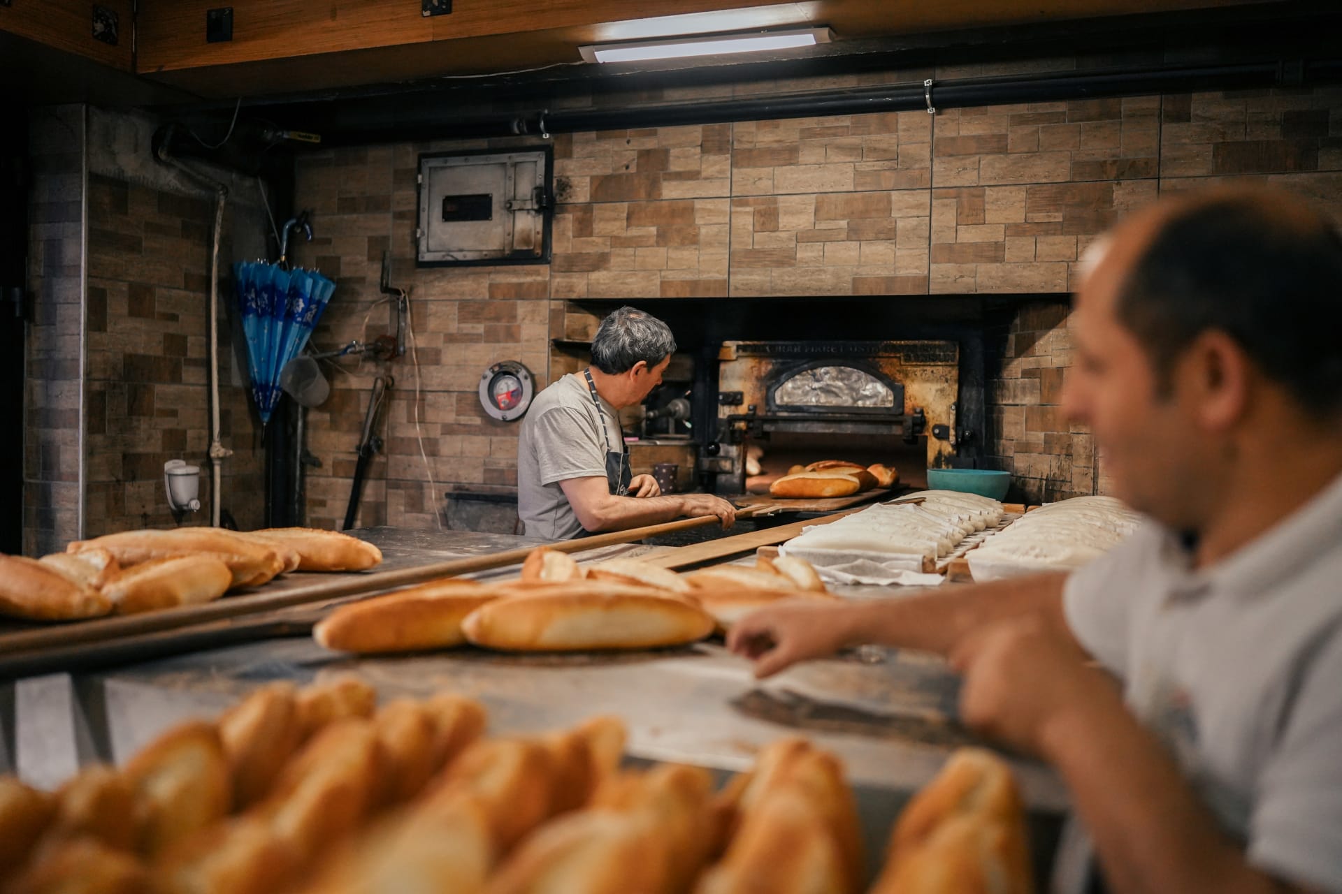 Two bakers at a stone oven, loaves on the counter.