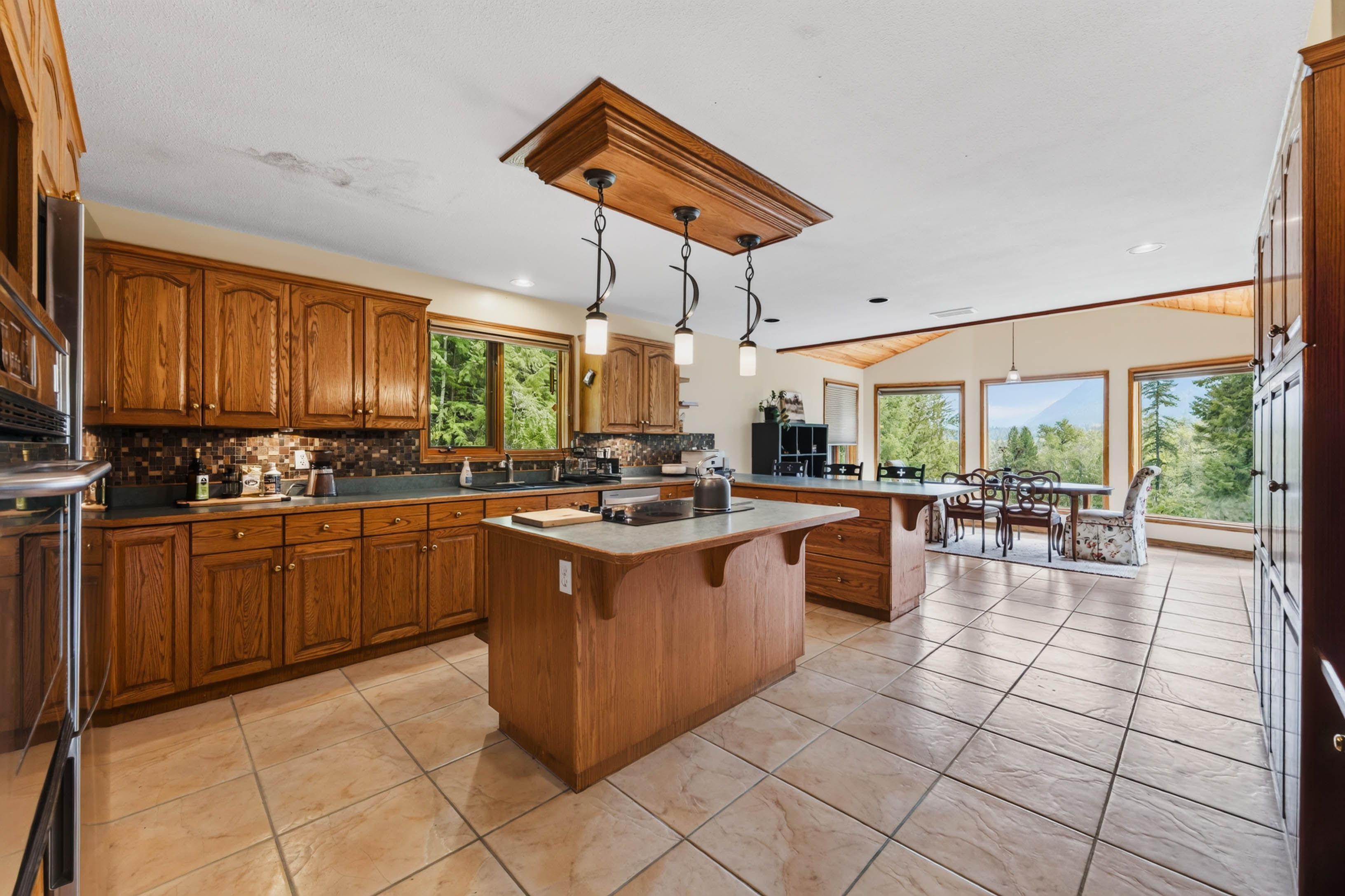 Open kitchen and dining area at 3360 Little Slocan South Road with wood cabinetry, central island, pendant lighting and large windows.