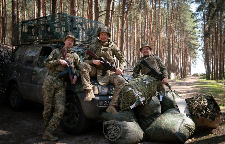 Троє бійців з автоматами біля пікапа, завантаженого камуфльованим спорядженням, у сосновому лісі