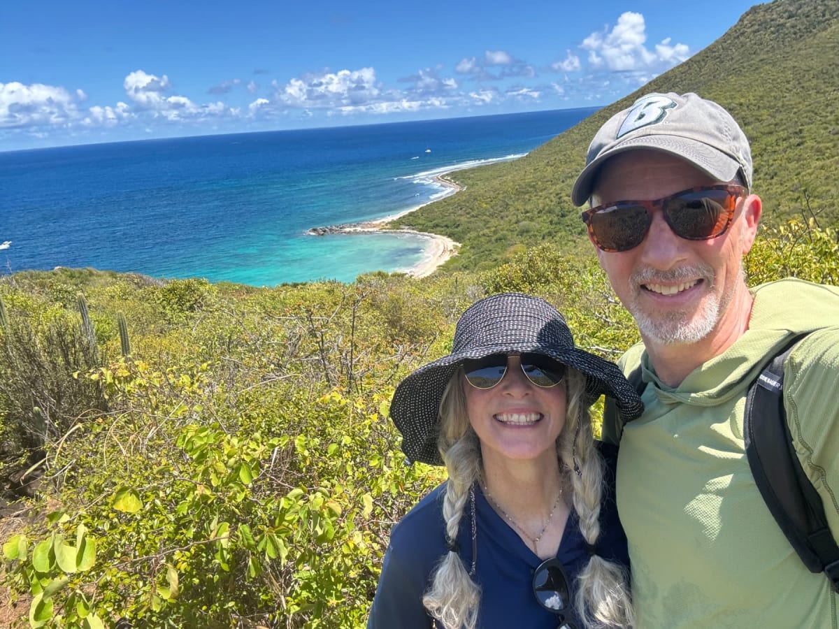 Two hikers smile at a scenic coastal overlook with turquoise waters and lush vegetation below.