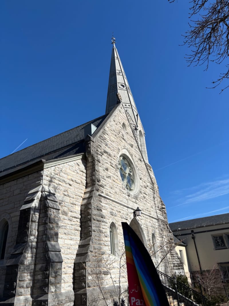 # Alt Text
Stone church with tall spire and circular window under clear blue sky, rainbow banner visible at entrance.
