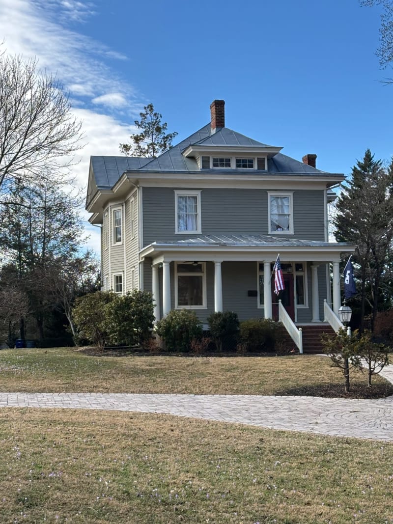 A classic two-story colonial house with gray siding, white columns, and metal roof on a sunny day.