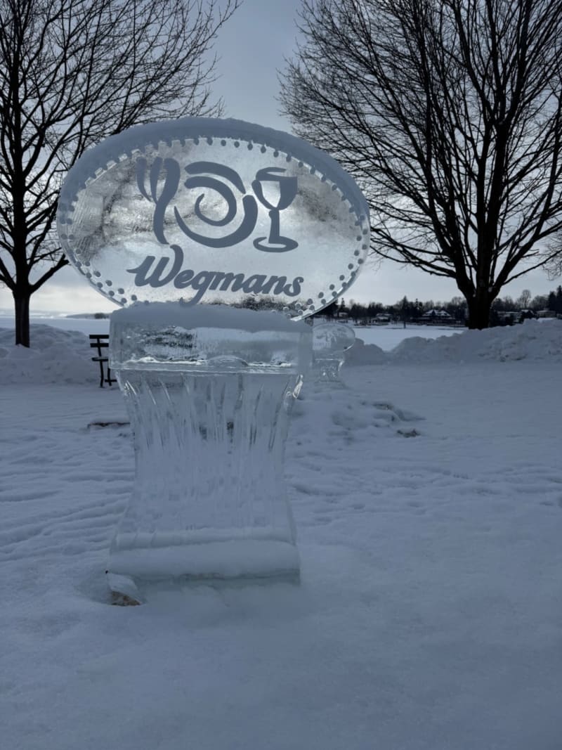 Frozen ice sculpture with "Wegmans" logo on a snowy landscape with bare trees and winter scenery.