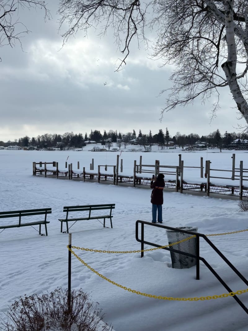 # Alt Text
A person stands on a frozen lake with wooden benches and yellow-chained barriers, waterfront homes visible across the ice.