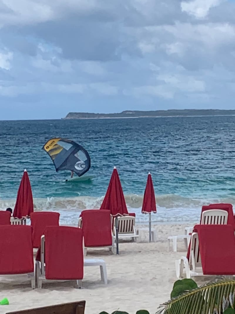 # Alt Text
Kiteboarder launches into the ocean near a beach resort with red umbrellas and loungers.