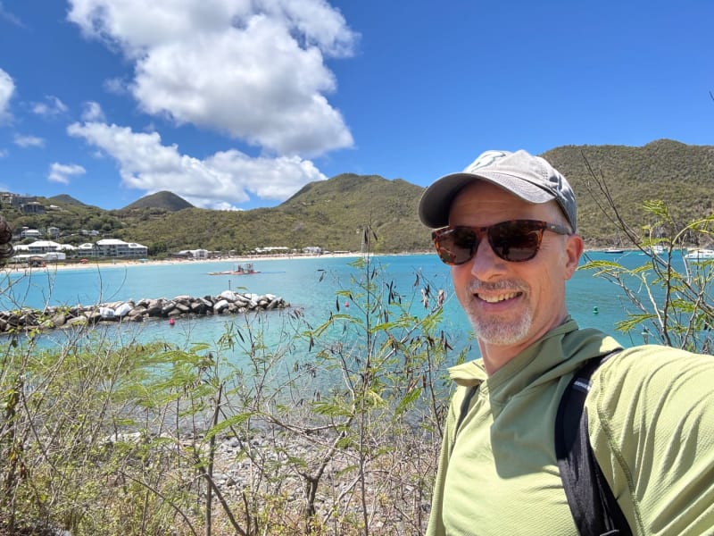 Man in sunglasses and cap smiling at a tropical beach cove with turquoise water and palm-covered hills.