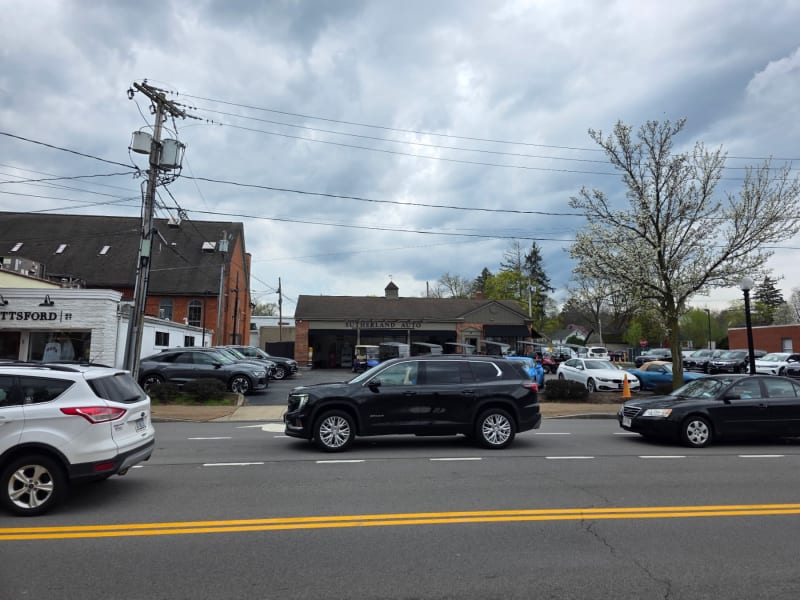 "Main street in a small New England town with parked cars, local businesses, and utility poles under a cloudy sky."
