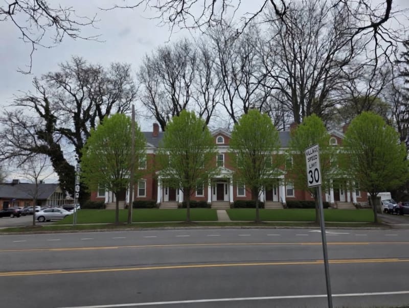 "Red brick colonial building with white columns and manicured lawn, framed by bare trees and a 30 mph village speed limit sign."