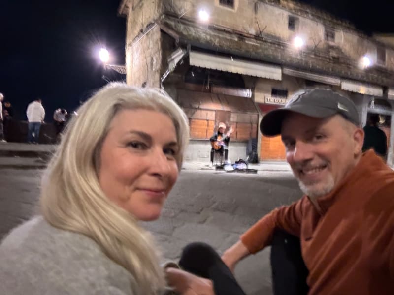 Two smiling people pose together in a historic town square at night with street musicians performing in the background.