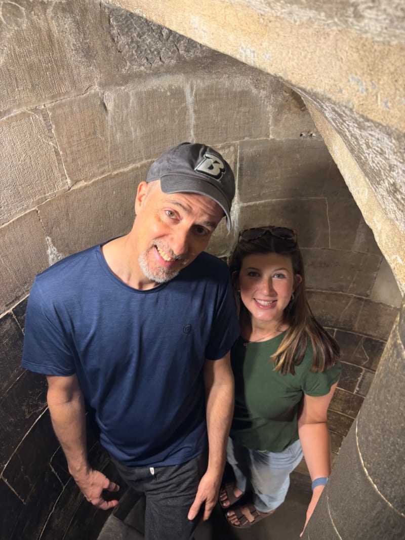 # Alt Text
Two people smile at the camera inside a narrow stone passageway or tunnel with arched ceiling.