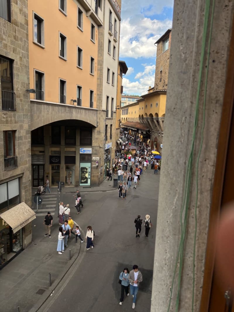 Busy medieval street in Florence with tourists, historic stone buildings, and yellow-ochre facades under blue sky.